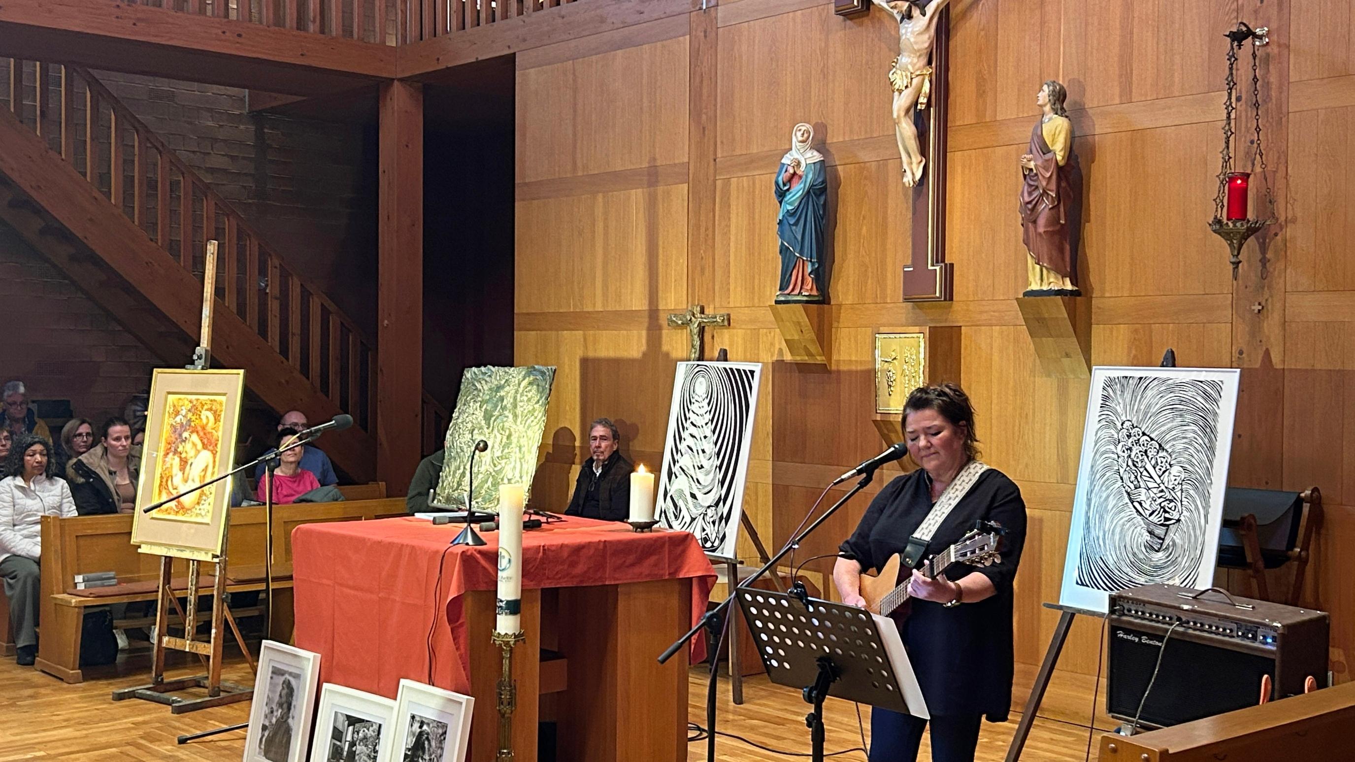 Innenraum einer Kirche mit holzverkleideten Wänden und einer erhöhten Empore im Hintergrund. Zentral an der Rückwand hängt ein großes Kruzifix, flankiert von zwei weiteren Heiligenfiguren. Vor dem Altarbereich stehen mehrere Staffeleien mit Kunstwerken, darunter farbige Gemälde und schwarz‑weiße Grafiken. Auf dem Boden vor dem Altar sind weitere gerahmte Bilder aufgereiht. Der Altar ist mit einem roten Tuch bedeckt, darauf stehen Kerzen und Mikrofone. Rechts im Vordergrund spielt eine Person Gitarre und singt in ein Mikrofon. Links und im Hintergrund sitzen Besucher in Kirchenbänken und verfolgen die Veranstaltung. Die Beleuchtung ist warm und lässt den Holzraum klar hervortreten.